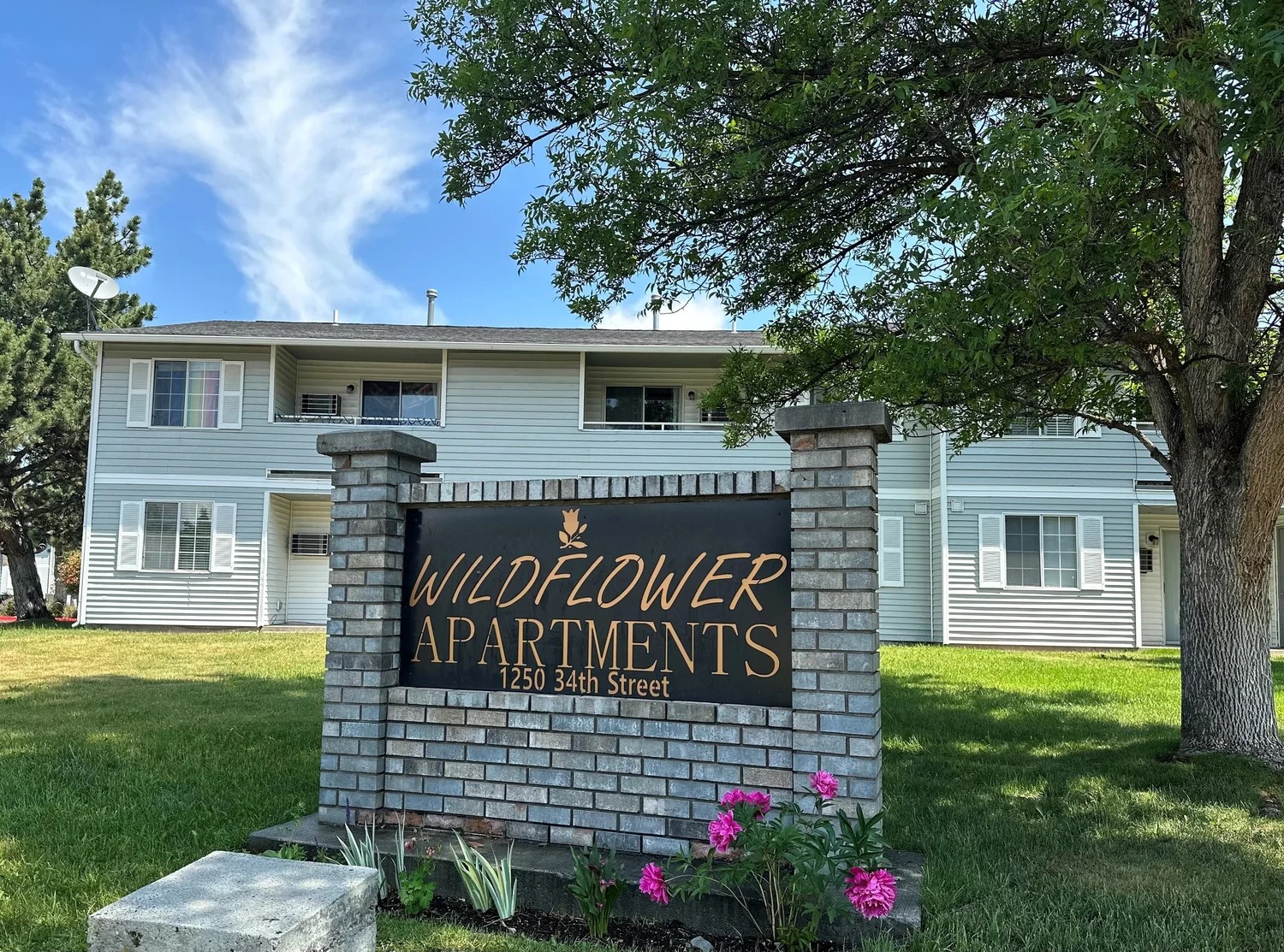 A black and gold sign over brick reads "Wildflower Apartments" in front of a two story apartment building